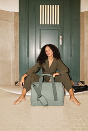 Frau sitzt vor einer grünen Tür mit einer großen, grünen Tasche im Vordergrund. Perspektive von vorne.