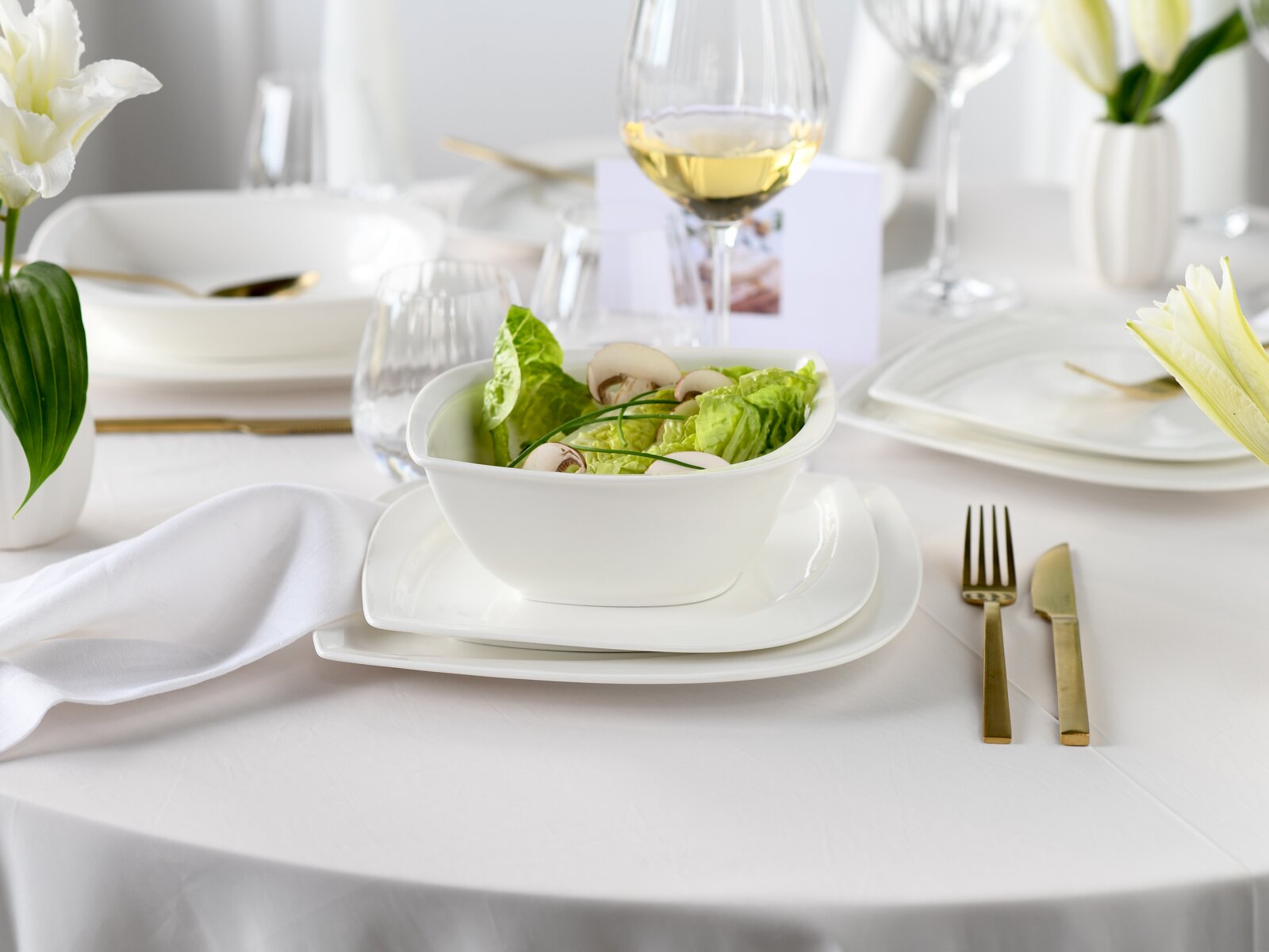 Weiße Porzellanschüssel mit grünem Salat und Champignons auf weißem Teller-Set, gedeckter Tisch mit goldfarbenem Besteck und Weißweinglas im Hintergrund, fotografiert aus leicht erhöhter Frontperspektive.