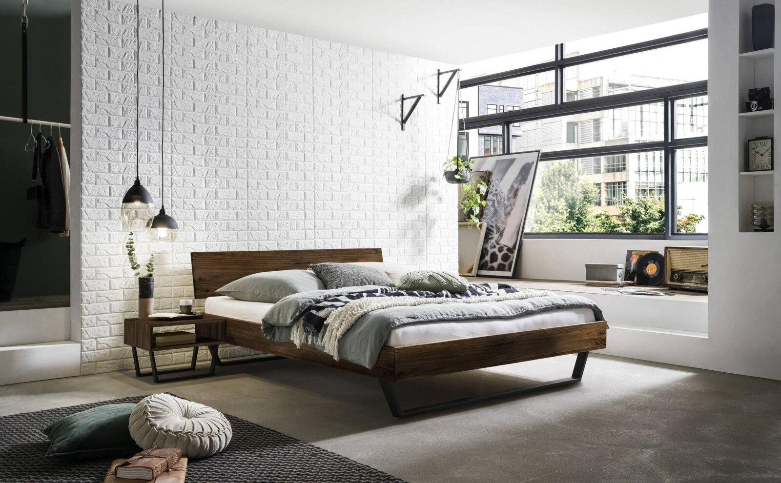 Moderne slaapkamer met een stijlvol houten bedframe, gezien vanaf de zijkant. Het bed is opgemaakt met grijs beddengoed en kussens, omgeven door een witte bakstenen muur en grote ramen die veel licht binnenlaten.