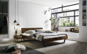 Moderne slaapkamer met een stijlvol houten bedframe, gezien vanaf de zijkant. Het bed is opgemaakt met grijs beddengoed en kussens, omgeven door een witte bakstenen muur en grote ramen die veel licht binnenlaten.