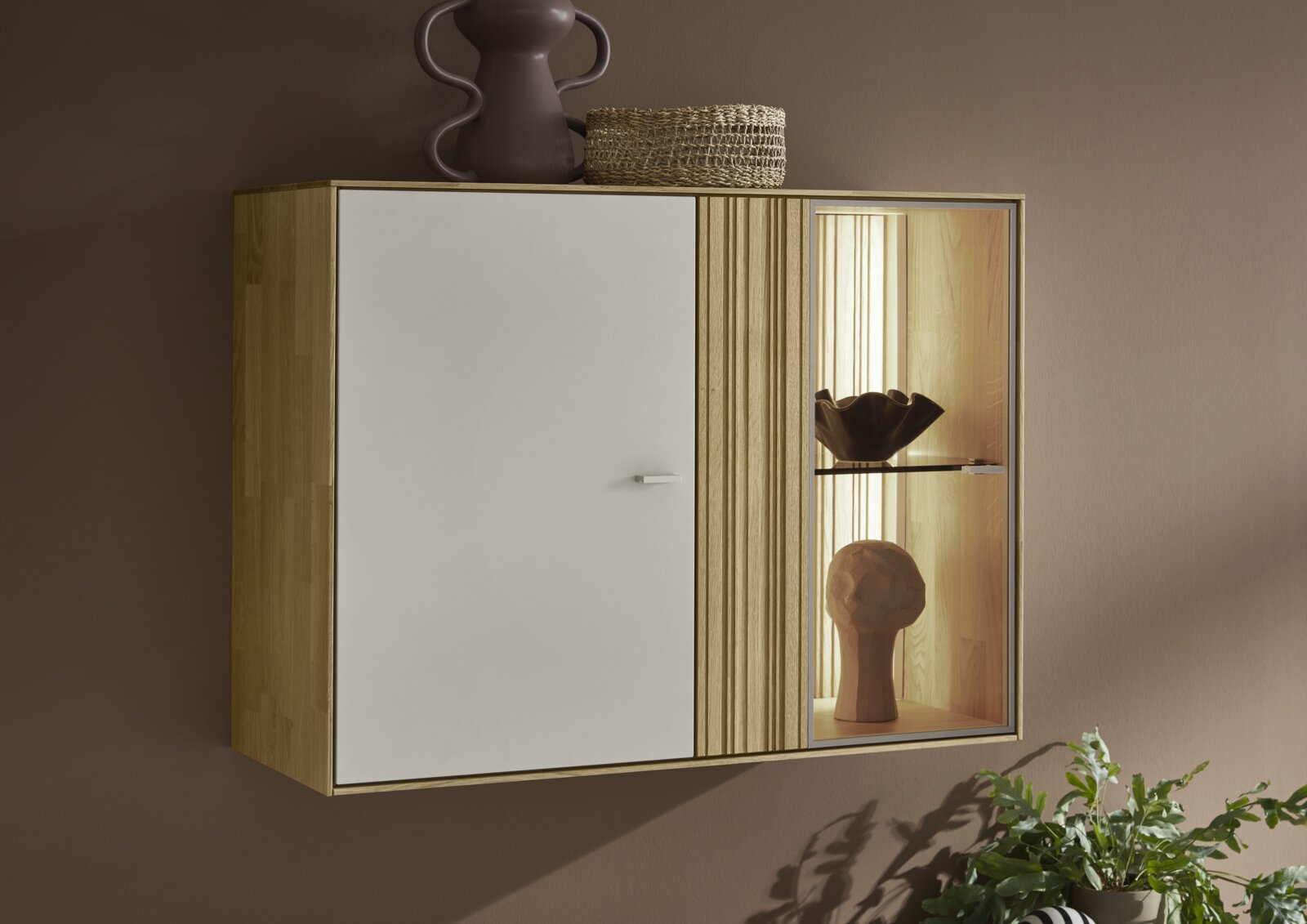 Wandhangende houten opbergkast met witte deur, verticale lamellen en verlichte glazen vitrine met decoratie, gefotografeerd in schuin vooraanzicht