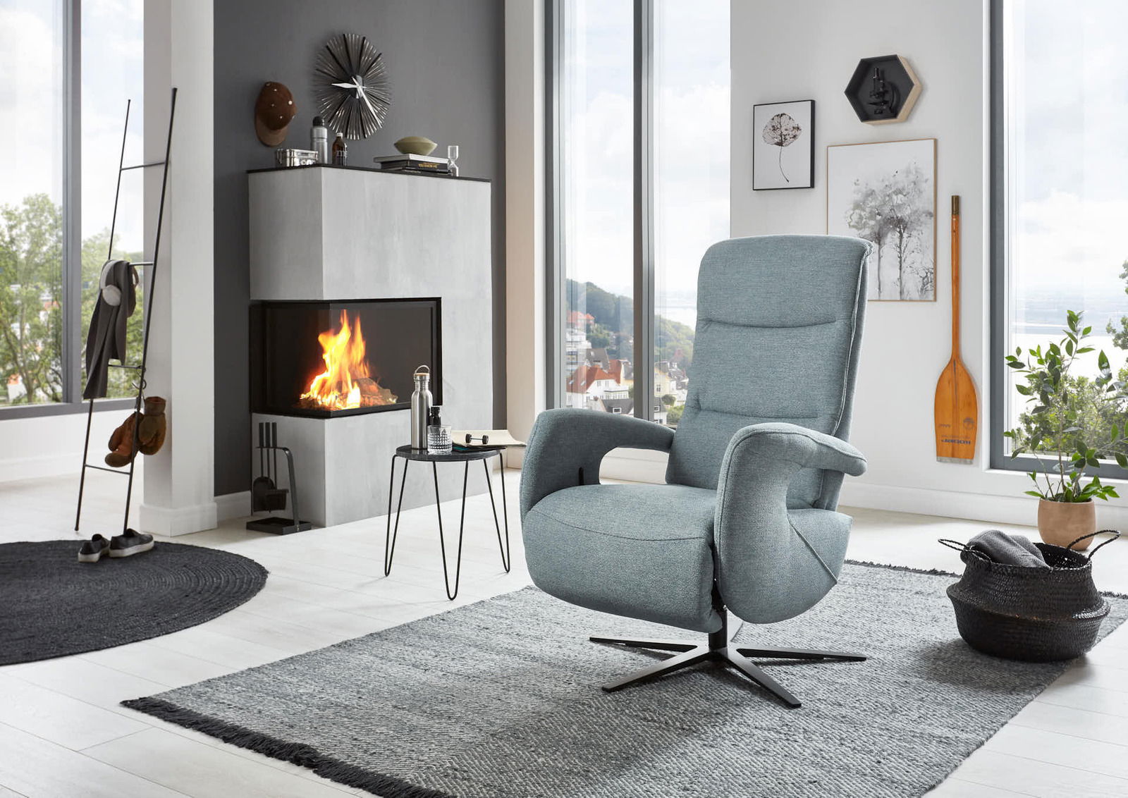 Moderner Relaxsessel in hellblauem Stoff, seitliche Perspektive, in einem stilvollen Wohnzimmer mit Kamin und großen Fenstern.