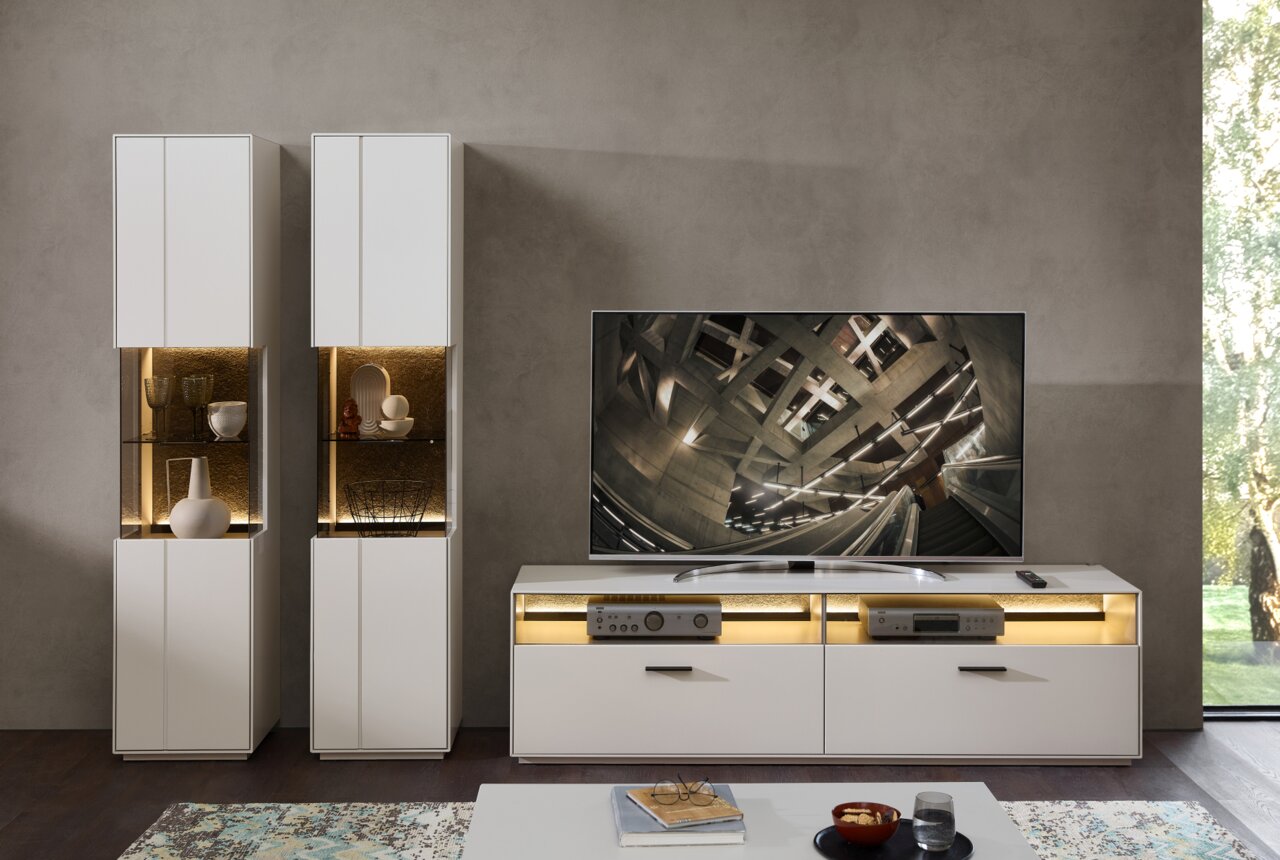 Een moderne woonkamer, gezien van voren, met twee witte vitrinekasten en een tv-meubel. De vitrinekasten zijn verlicht met decoratieve voorwerpen.