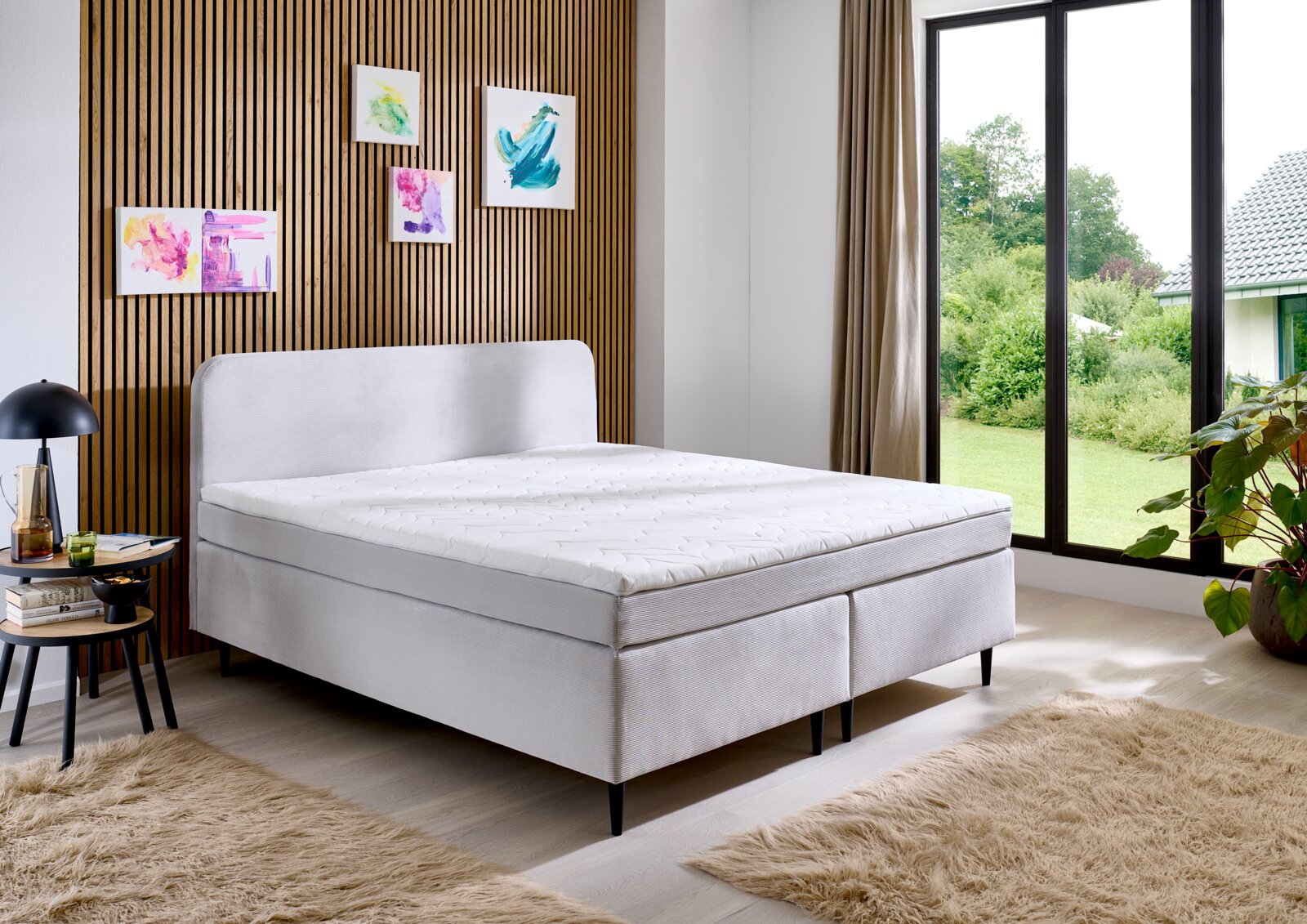 Modern boxspringbed in een stijlvolle slaapkamer met houten lambrisering en grote ramen, zijaanzicht.