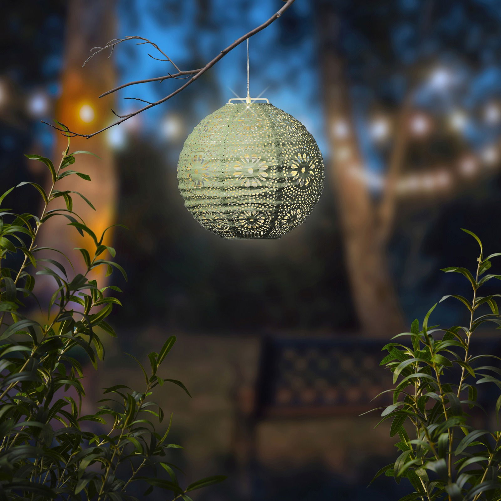 Solar Laterne in Kugelform, die an einem Ast hängt, mit dekorativem Muster und warmem Licht, im Hintergrund ein unscharfer Garten bei Dämmerung.