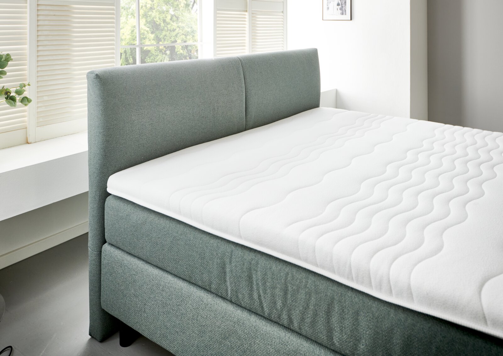 Boxspringbett BX 180/200 in seitlicher Perspektive mit grauem Stoffbezug und weißer Matratze in einem hellen Raum mit Fenster.