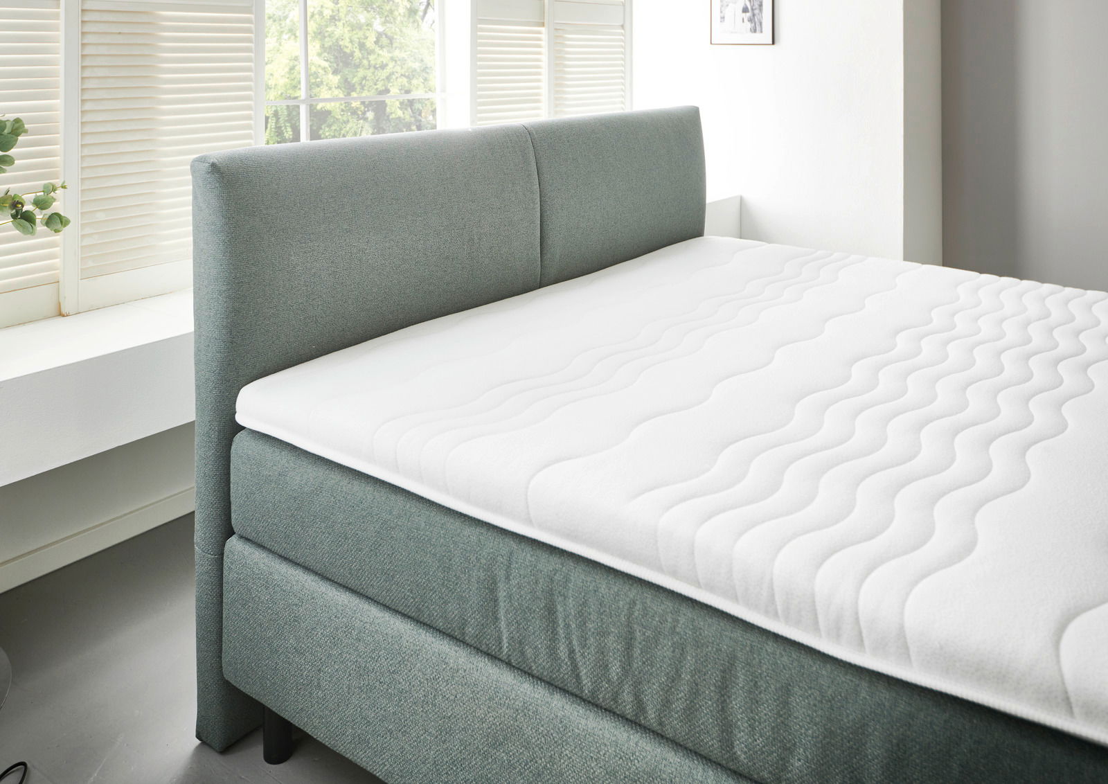 Boxspringbett BX 180/200 in seitlicher Perspektive mit grauem Stoffbezug und weißer Matratze in einem hellen Raum mit Fenster.