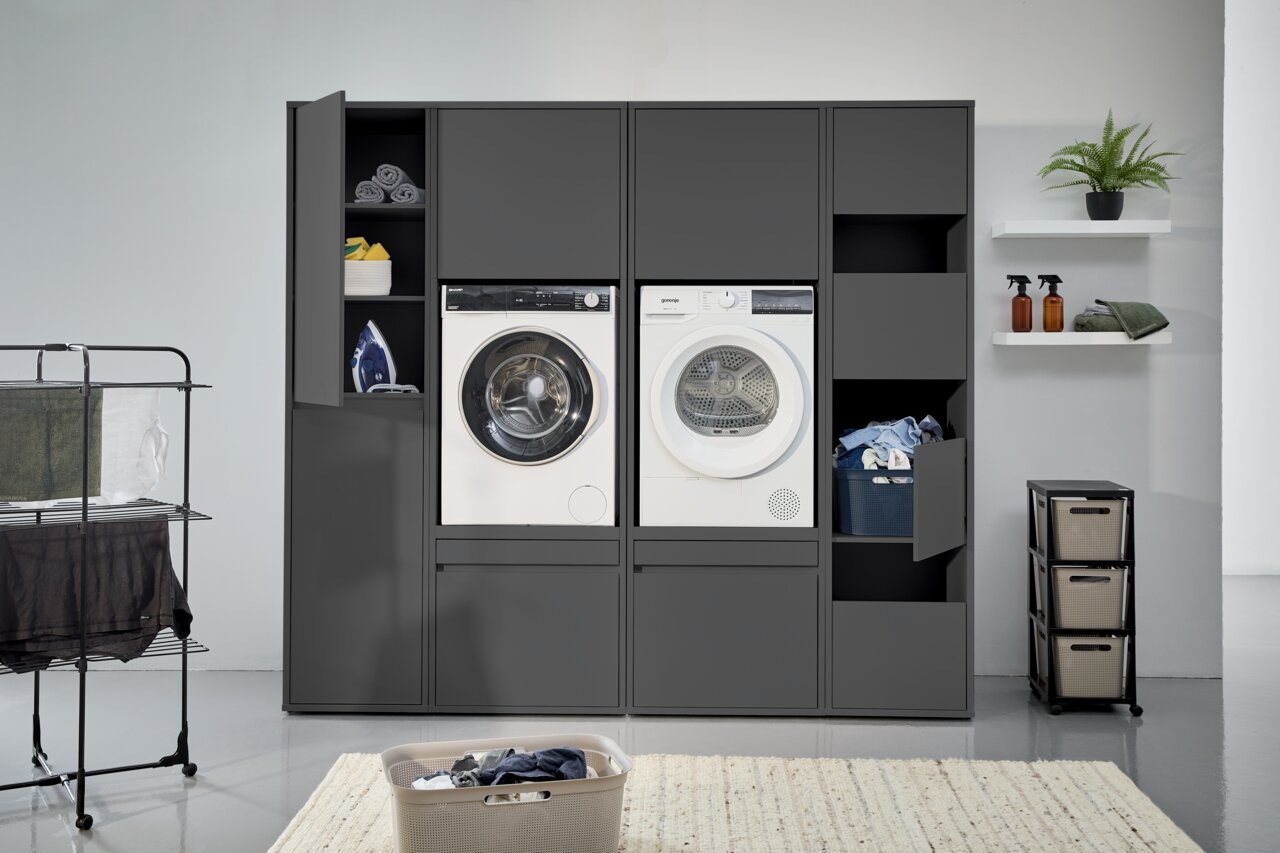 XORA Doppel-Waschmaschinenschrank LIV WASHTOWER Frontansicht eines Doppel-Waschmaschinenschranks mit zwei Waschmaschinen, offenen Fächern mit Handtüchern und einem Bügeleisen, sowie einem Wäschekorb und Regalen im Hintergrund.