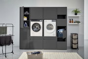 Vooraanzicht van een dubbele wasmachine-kast met twee wasmachines, open vakken met handdoeken en een strijkijzer, evenals een wasmand en planken op de achtergrond.
