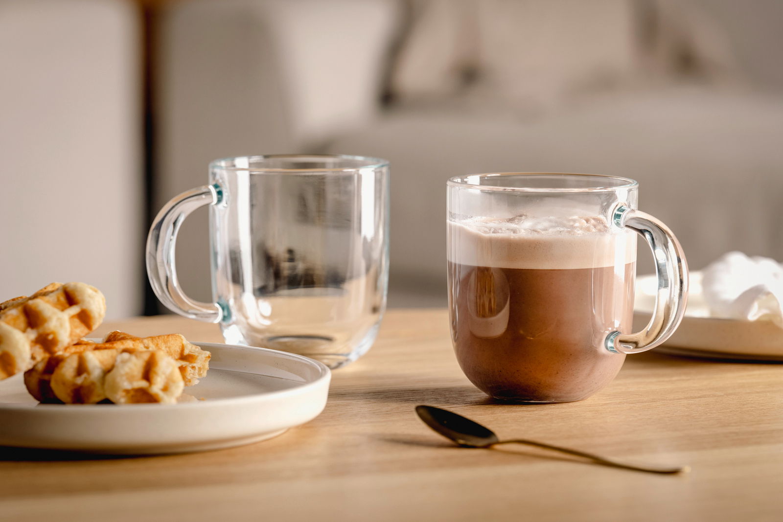 Zwei transparente Tassen auf einem Holztisch, eine gefüllt mit heißer Schokolade, die andere leer, mit einem Teller Waffeln und einem goldenen Löffel im Vordergrund. Seitliche Perspektive.