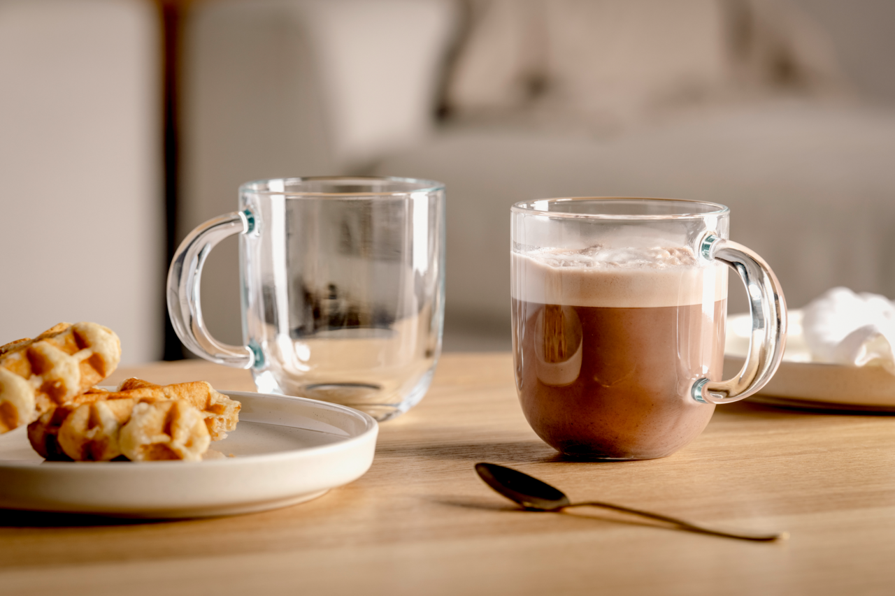 Zwei transparente Tassen auf einem Holztisch, eine gefüllt mit heißer Schokolade, die andere leer, mit einem Teller Waffeln und einem goldenen Löffel im Vordergrund. Seitliche Perspektive.