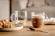 Twee doorzichtige kopjes op een houten tafel, de ene gevuld met warme chocolademelk, de andere leeg, met op de voorgrond een bord wafels en een gouden lepel. Zijaanzicht.