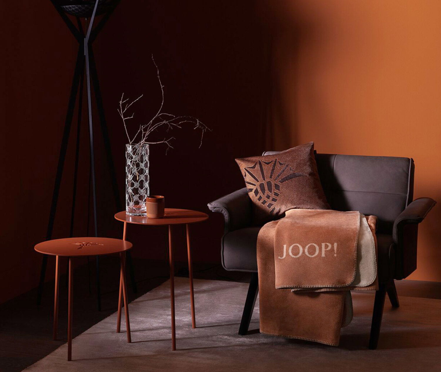 Wohnbereich mit dunklem Sessel, dekorativen Kissen, braunen Beistelltischen und einer hell-orangen Decke vor einem Hintergrund in sattem Orange.