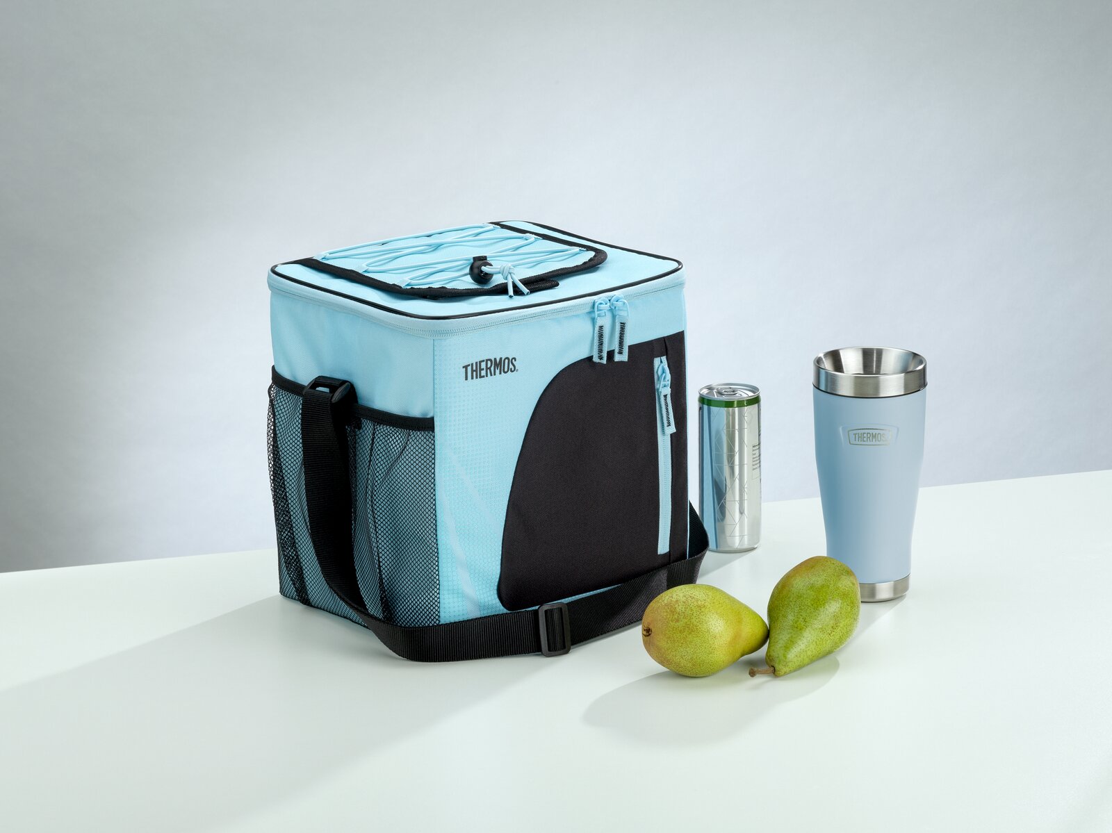 Hellblaue THERMOS-Kühltasche mit schwarzem Schultergurt und Außentaschen, aus schräger Vorderansicht neben Thermobecher, Getränkedose und zwei Birnen präsentiert