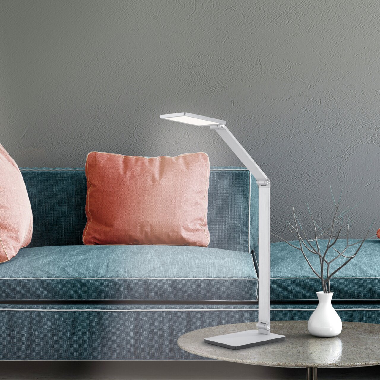 Moderne tafellamp van aluminium, gezien vanaf de zijkant, naast een bank met roze kussens en een tafeltje met een vaas.