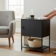 Schwarzer, minimalistischer Beistelltisch/Nachttisch aus Metall mit perforierter Klapptür und Ablagefach, neben grauem Sessel im Wohnzimmer; perspektivisch schräg von vorn rechts aufgenommen.