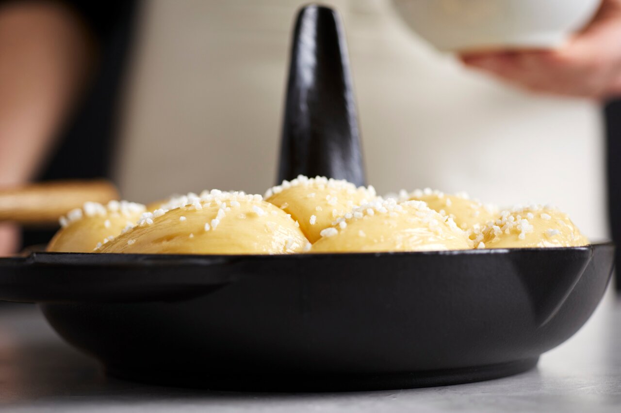 Close-up van een zwarte STAUB-kippenbraadpan met deegballetjes die met suiker zijn bestrooid. De braadpan heeft een centrale kegel en is vanuit zijaanzicht gefotografeerd.