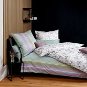 Schlafzimmer-Setting mit Bett in gestreifter Bettwäsche in Pastellgrün und Flieder sowie weißer Bettdecke mit zartem Blumenmuster, Kissen in Grün und Mauve; Ansicht schräg von vorne links.