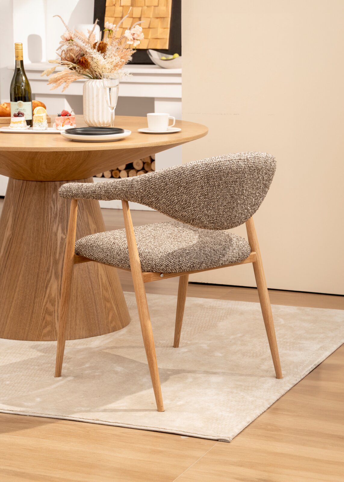 Gepolsterter Esszimmerstuhl mit grauem Strukturstoff und Holzbeinen am runden Holztisch auf hellem Teppich, perspektivisch in Seitenansicht von schräg vorne.