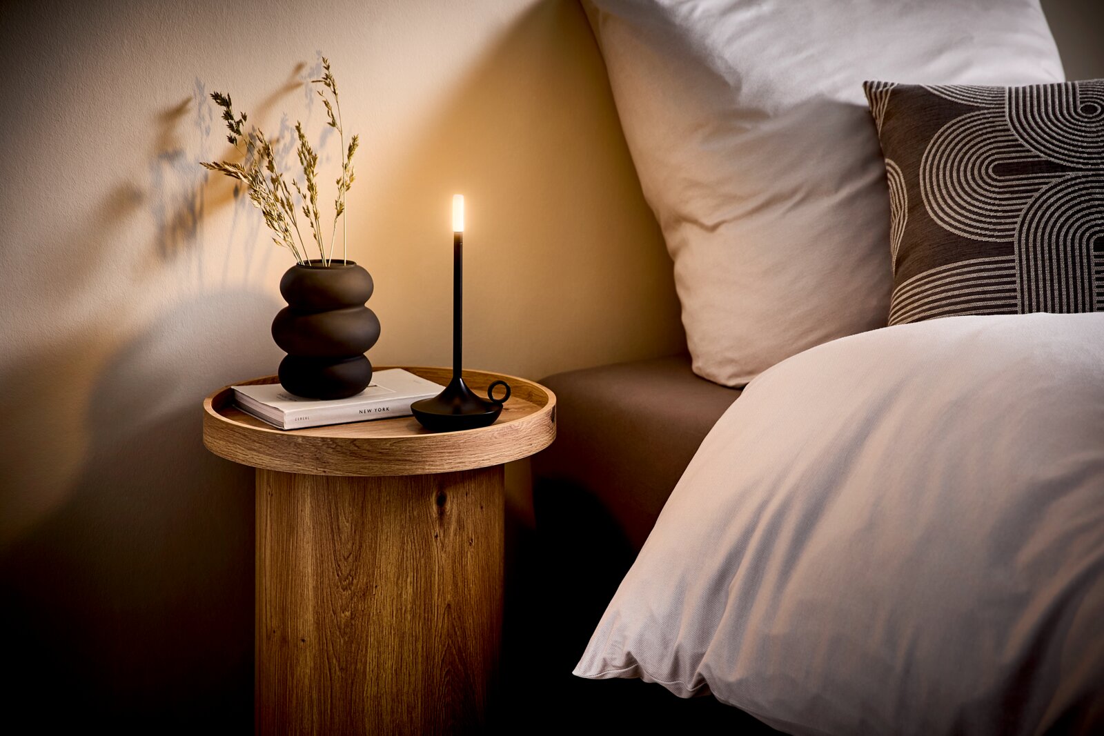 Stilvoller runder Nachttisch aus Holz neben einem Bett, mit schwarzer Vase, Buch und schlanker LED-Tischlampe; Perspektive: seitliche Ansicht auf Augenhöhe