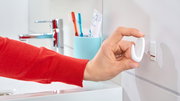 Hand befestigt einen weißen tesa Powerstrips Haken Small an einer Badezimmerwand, seitliche Perspektive.