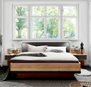 Modern houten futonbed met gestoffeerd hoofdeinde, geflankeerd door nachtkastjes, in een lichte kamer met grote ramen, vooraanzicht
