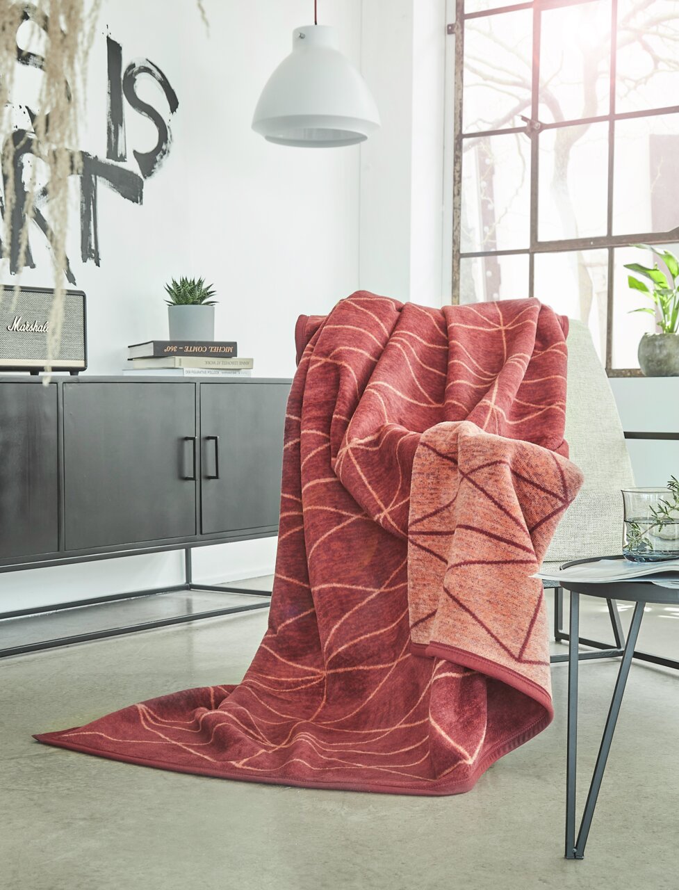 IBENA Decke 150x200cm BUGATTI EX Rote Jacquard-Decke 150x200 cm mit Blattmuster, drapiert über einem Sessel in einem modernen Wohnzimmer mit Blick auf Fenster und Dekoration.