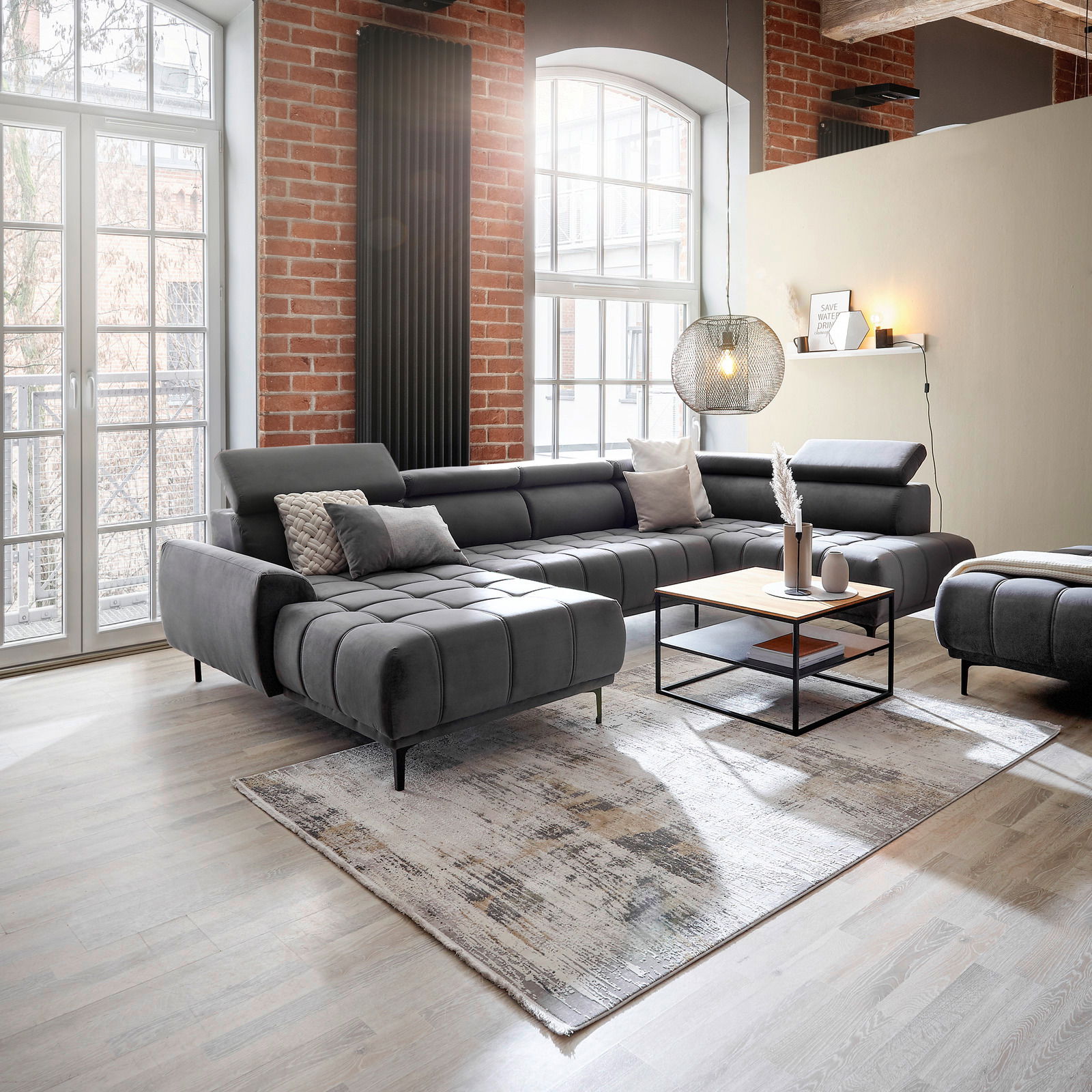 Moderne graue Wohnlandschaft in einem hellen Wohnzimmer mit Backsteinwand und großen Fenstern, seitliche Perspektive.