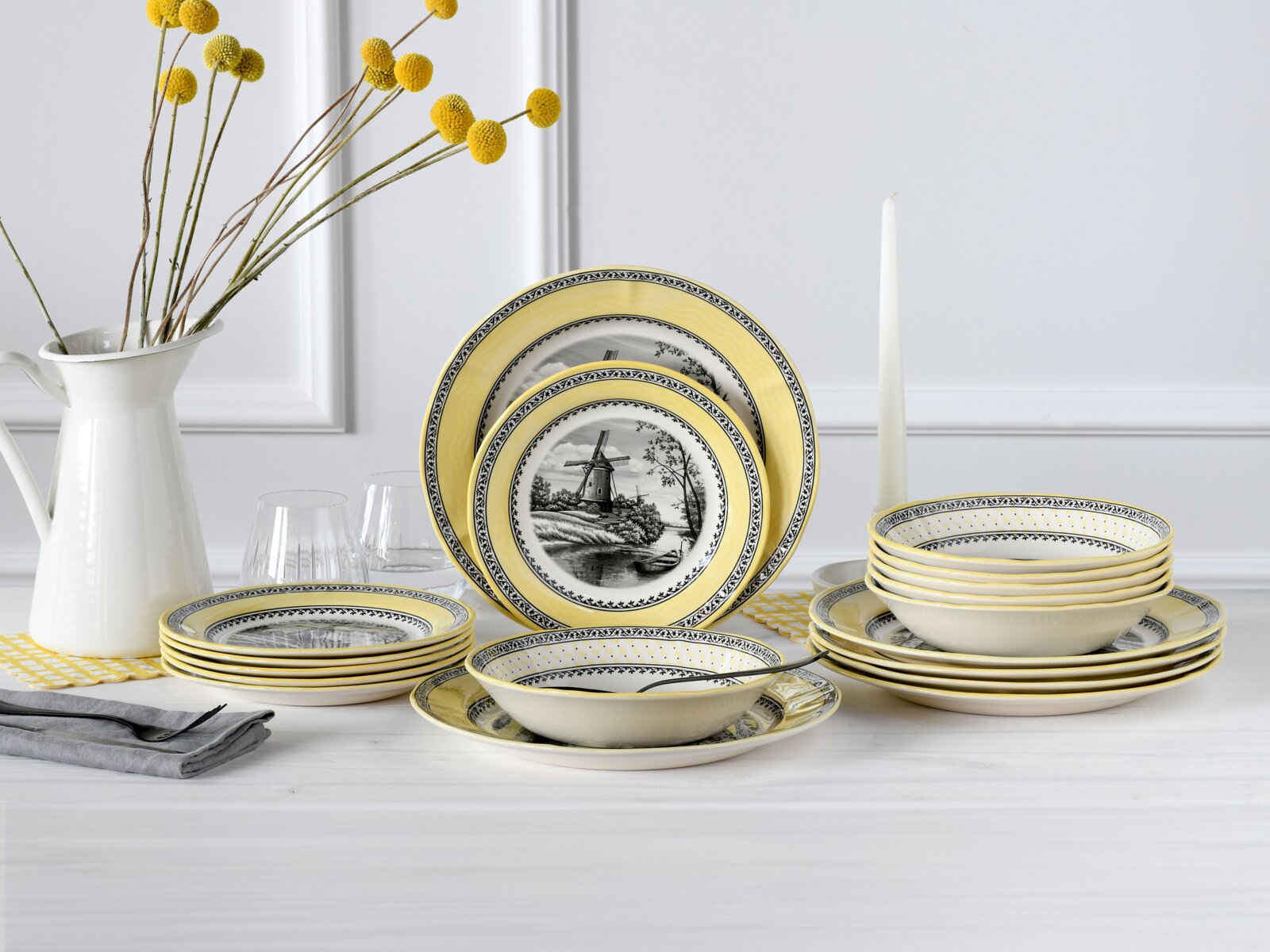 creaTable Tellerset 18-tlg WINDMÜHLE Gelb-weißes Keramik-Geschirrset mit schwarzem Windmühlenmotiv und gemustertem Rand, Teller und Schalen auf einem Tisch arrangiert; Perspektive: frontale Ansicht auf Tischhöhe.