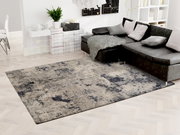 Moderner Designer-Teppich in einem stilvollen Wohnzimmer, mit einer grauen und blauen Musterung, neben einem dunklen Ecksofa. Perspektive von oben.