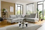 Moderne lichtgrijze L-vormige hoekbank met chaise longue en zwarte metalen poten in een lichte woonkamer, gezien van voren en iets van opzij.