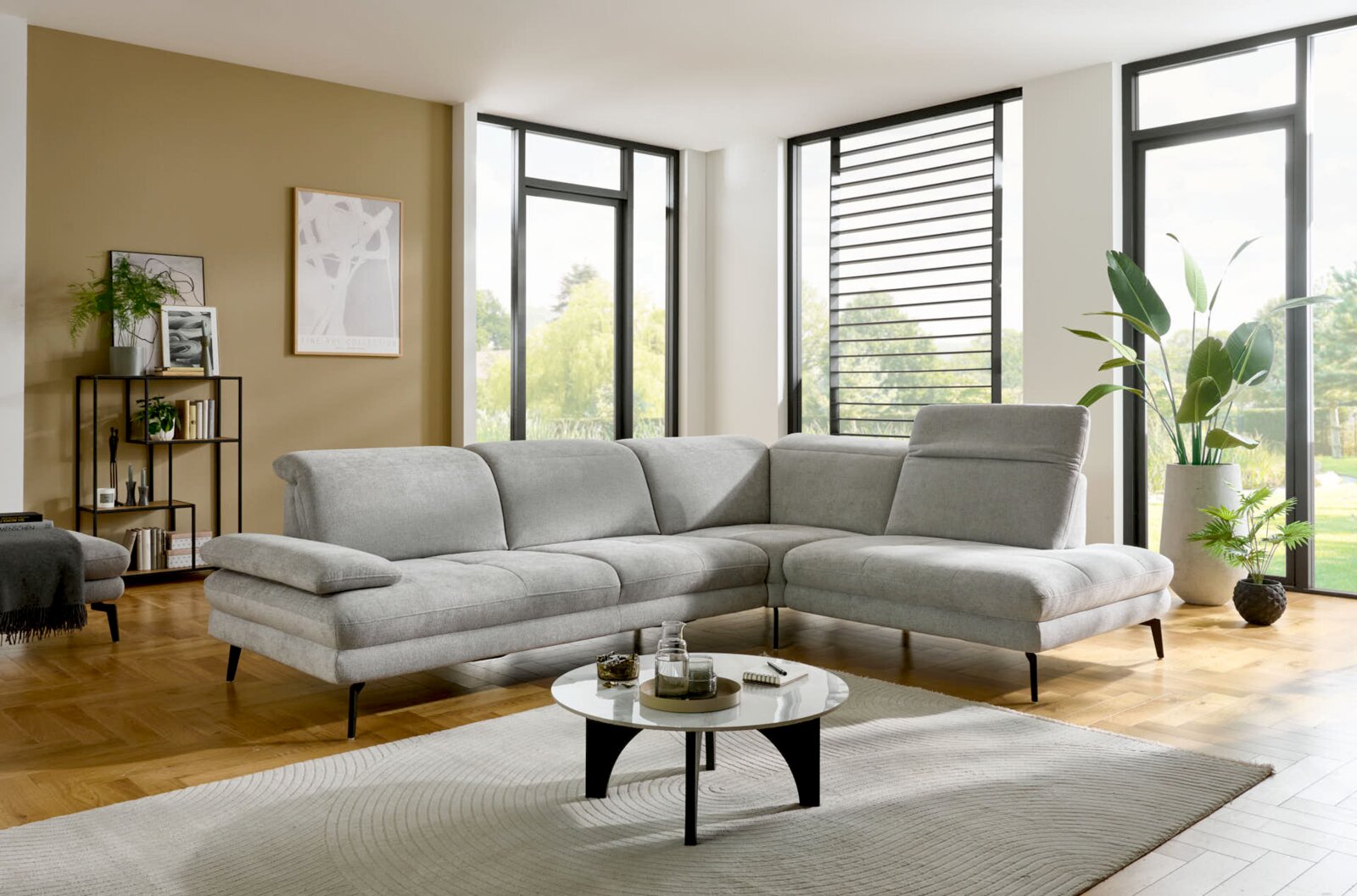 Moderne lichtgrijze L-vormige hoekbank met chaise longue en zwarte metalen poten in een lichte woonkamer, gezien van voren en iets van opzij.