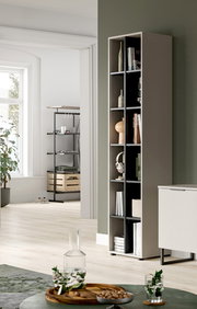 Schmales, hohes Regal in Taupe mit offenen Fächern und Deko, in modernem Wohnraum an grüner Wand, perspektivisch in leichter Seitenansicht von vorn rechts