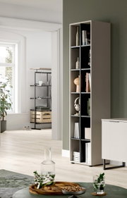 Schmales, hohes Regal in Taupe mit offenen Fächern und Deko, in modernem Wohnraum an grüner Wand, perspektivisch in leichter Seitenansicht von vorn rechts