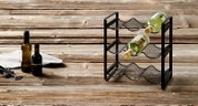 Flessenrek van zwart metaal met drie niveaus, zijaanzicht, op een rustieke houten tafel met twee wijnflessen.
