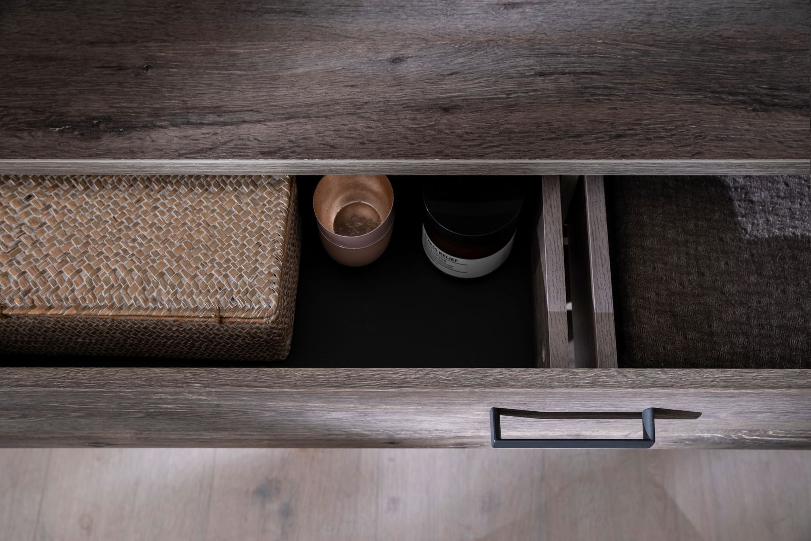 Nahaufnahme einer geöffneten Schublade eines Sideboards mit dunkler Holzoptik, gefüllt mit einem geflochtenen Korb, einer Kerze und einem Glasbehälter. Perspektive von oben.