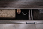 Nahaufnahme einer geöffneten Schublade eines Sideboards mit dunkler Holzoptik, gefüllt mit einem geflochtenen Korb, einer Kerze und einem Glasbehälter. Perspektive von oben.