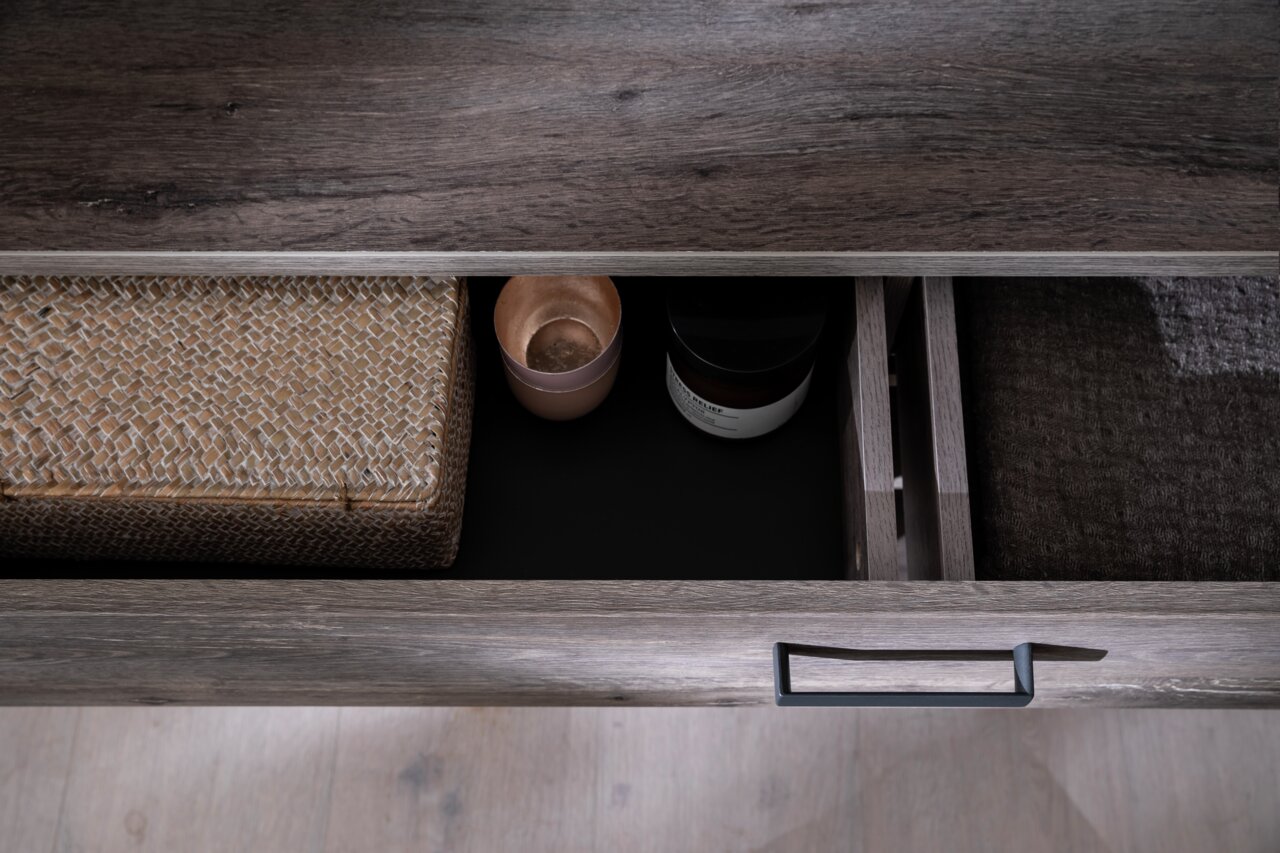 Close-up van een geopende lade van een dressoir met een donkere houtlook, gevuld met een gevlochten mand, een kaars en een glazen pot. Aanzicht van bovenaf.