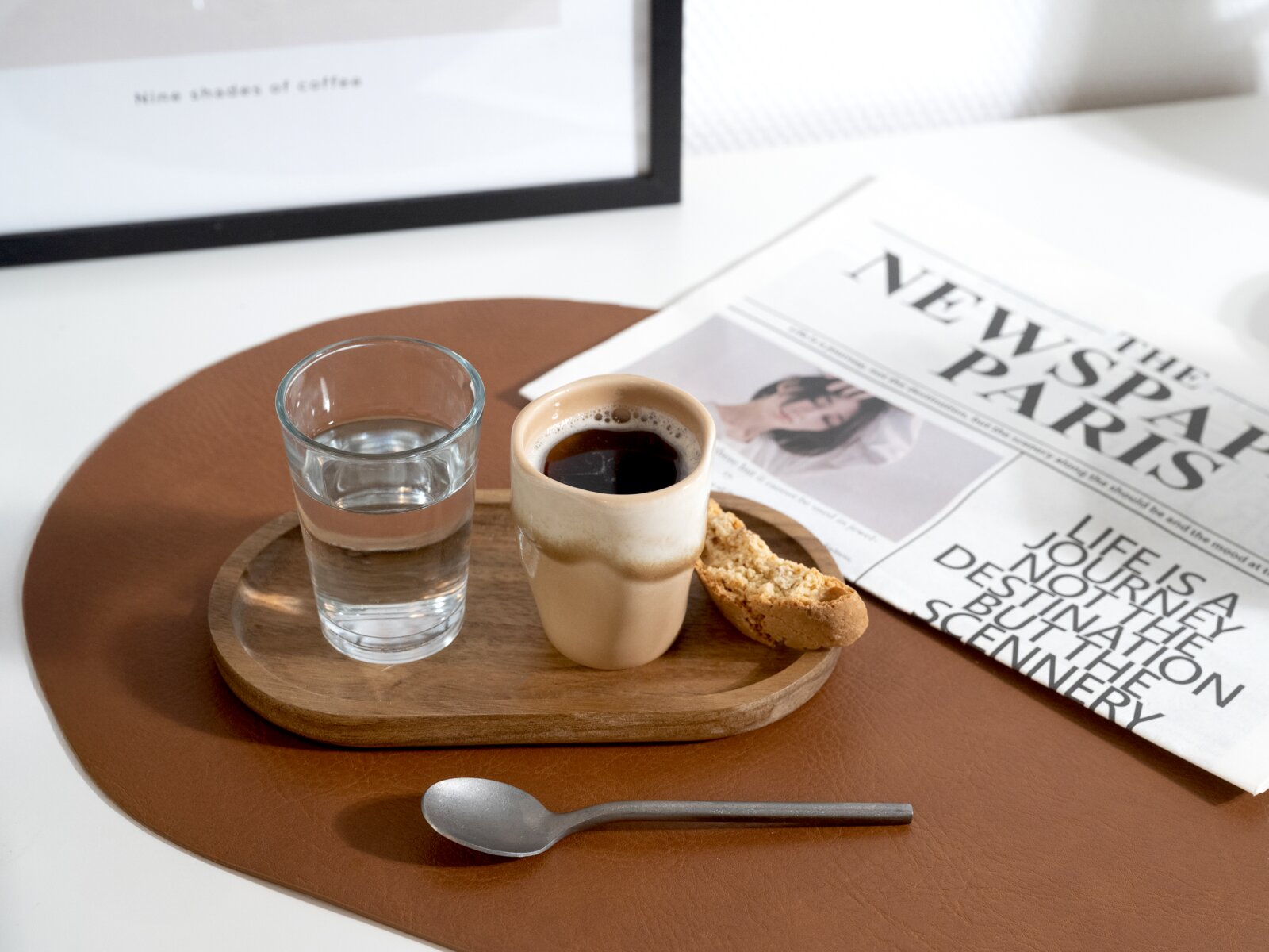 Holztablett mit Espresso in beigem Becher, Wasserglas und Biscotti auf braunem Leder-Set, daneben Zeitung und Löffel – Perspektive: leicht von oben (Top-Down)