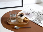 Holztablett mit Espresso in beigem Becher, Wasserglas und Biscotti auf braunem Leder-Set, daneben Zeitung und Löffel – Perspektive: leicht von oben (Top-Down)