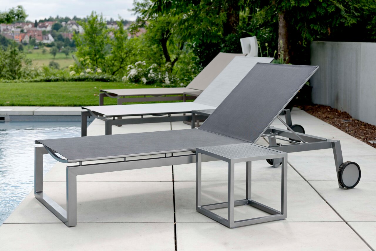 Moderne Rollenliege aus Metall mit verstellbarer Rückenlehne, seitliche Perspektive am Pool mit grüner Landschaft im Hintergrund.