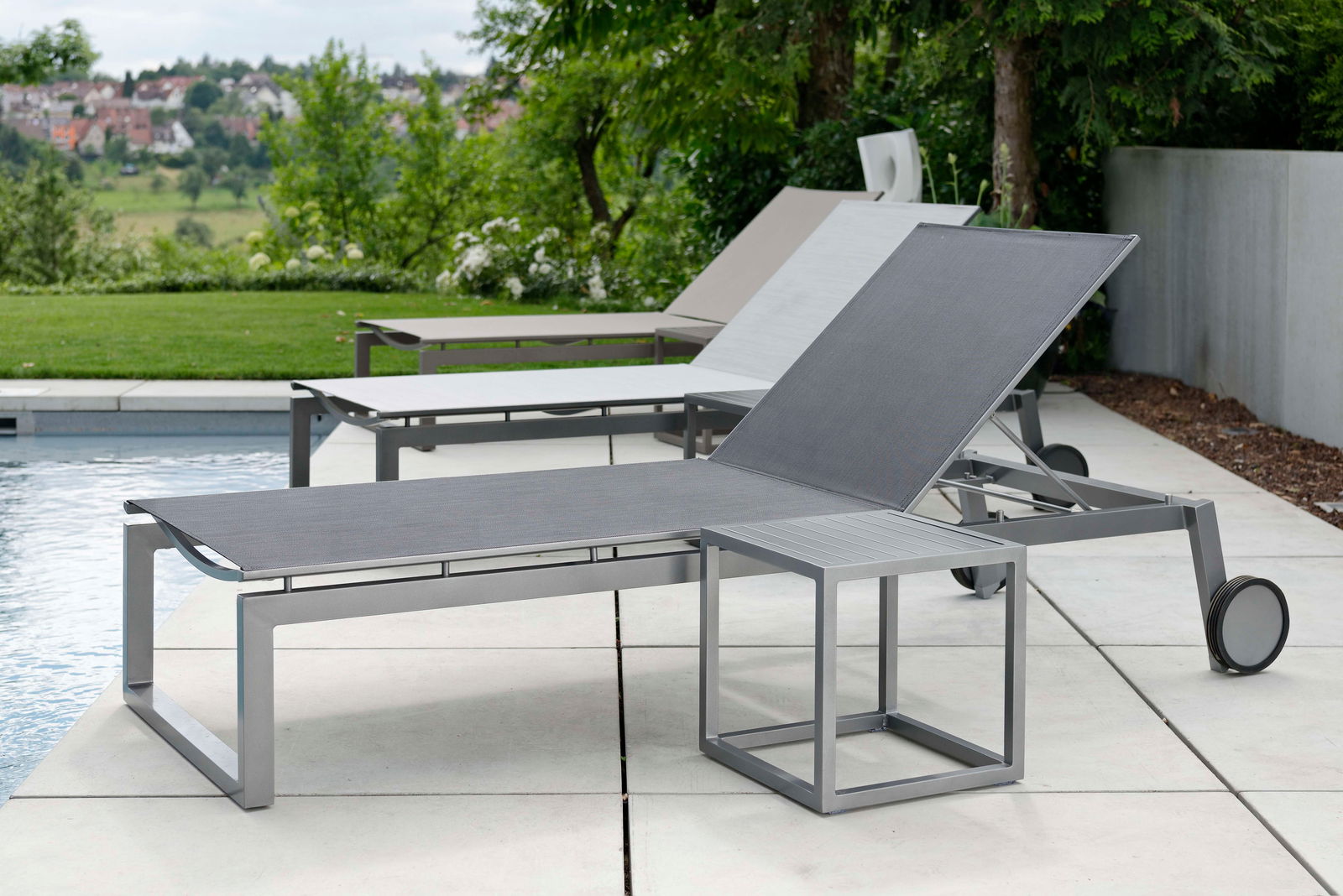 Moderne Rollenliege aus Metall mit verstellbarer Rückenlehne, seitliche Perspektive am Pool mit grüner Landschaft im Hintergrund.