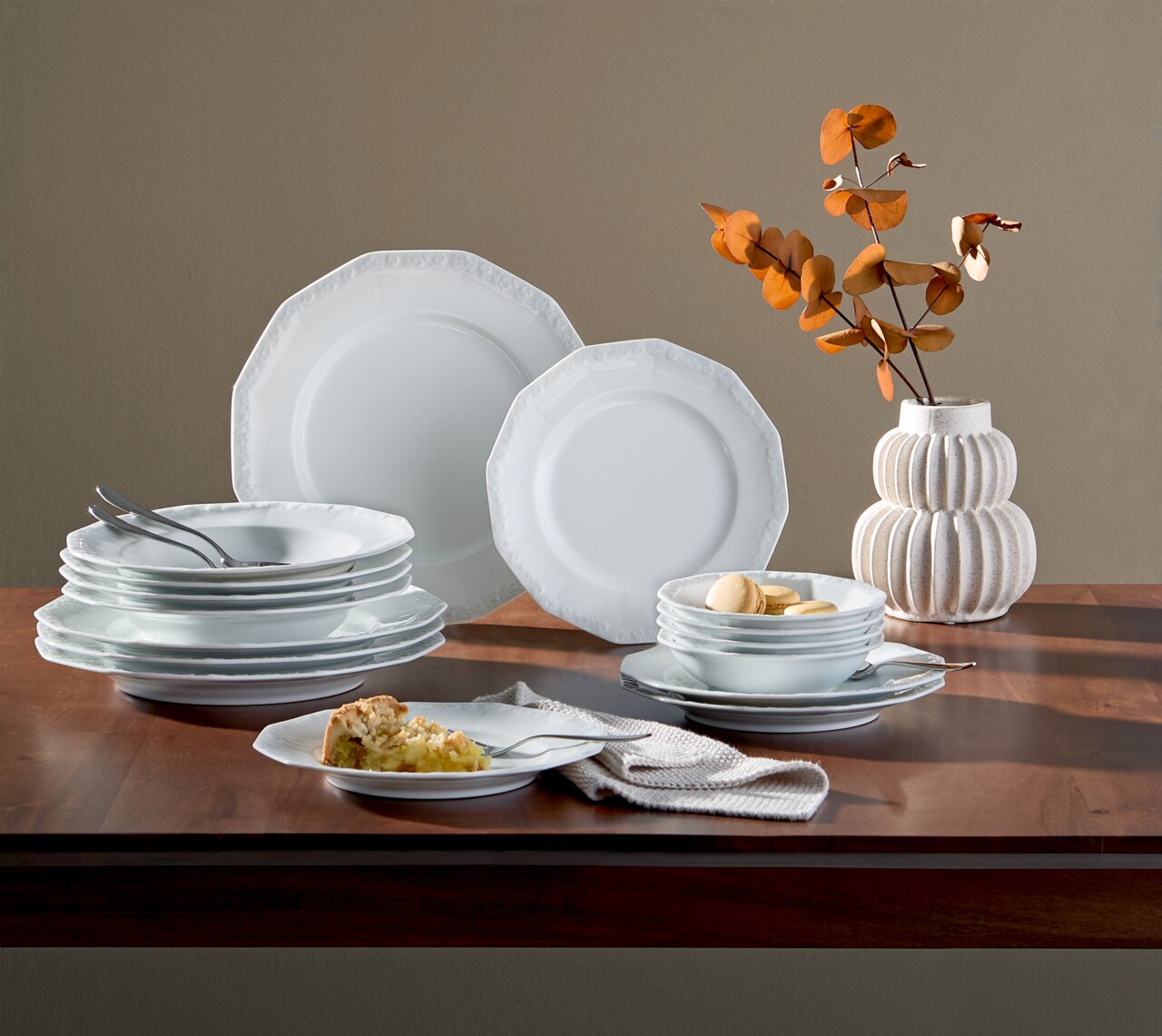 Kombiservice Maria Weiß 16-tlg auf einem Tisch, seitliche Perspektive, bestehend aus weißen Tellern und Schalen mit dezentem Muster, dekoriert mit einem Strauß getrockneter Blätter in einer Vase.