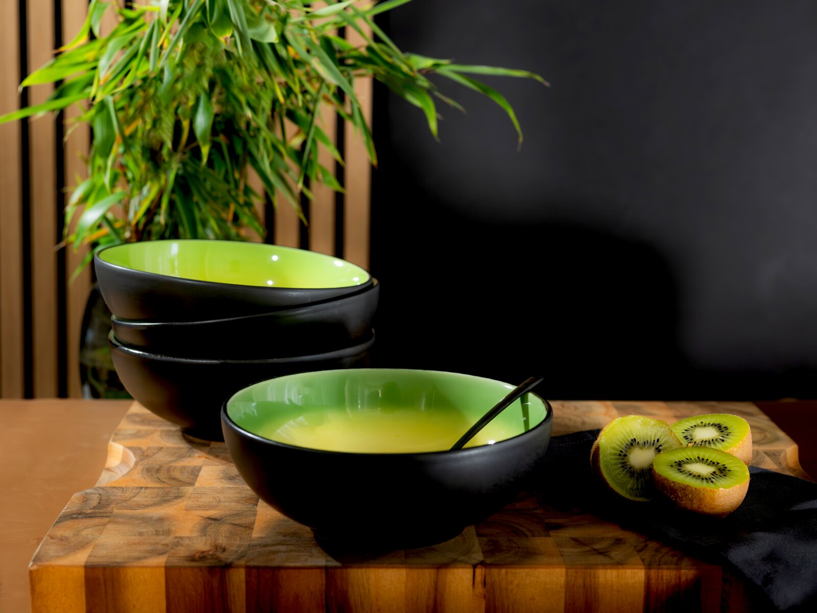 Schwarze Schalen mit leuchtend grüner Innenseite, aus schräger Vorderansicht auf einem Holzbrett mit Löffel und frischen Kiwis inszeniert
