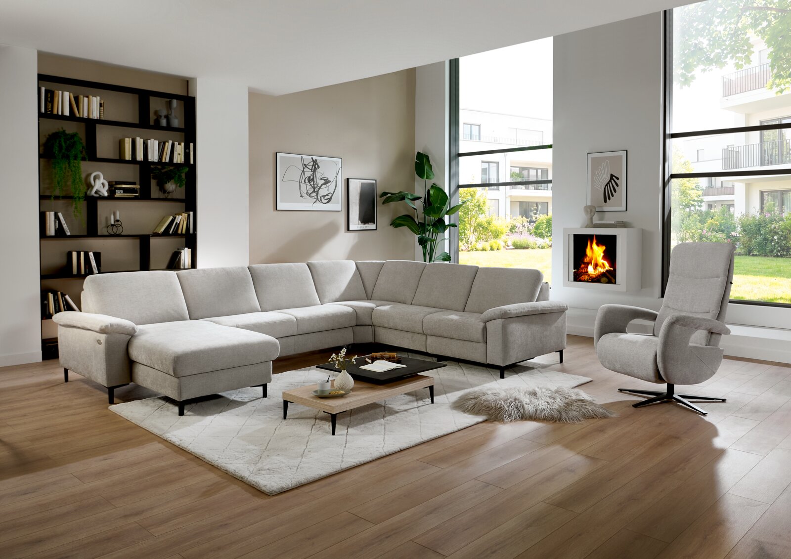 Modernes hellgraues L-förmiges Ecksofa mit Chaiselongue in einem hellen Wohnzimmer mit Couchtisch, Teppich und Sessel; Perspektive: frontale Weitwinkelansicht leicht von links.