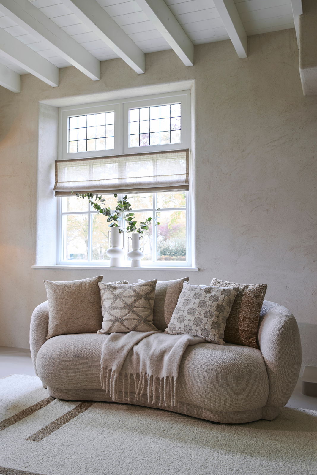 Gemütliches Wohnzimmer mit einem beigen Sofa, dekoriert mit mehreren Kissen in neutralen Tönen, vor einem Fenster mit Blick auf den Garten. Frontalansicht.