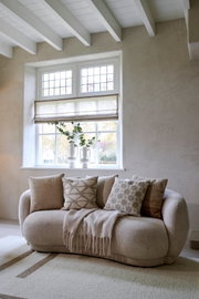 Gemütliches Wohnzimmer mit einem beigen Sofa, dekoriert mit mehreren Kissen in neutralen Tönen, vor einem Fenster mit Blick auf den Garten. Frontalansicht.