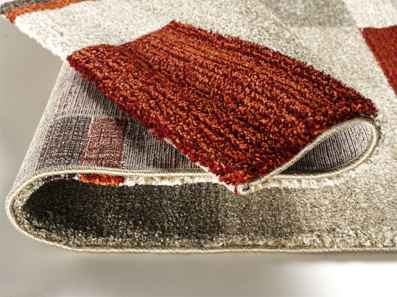 Nahaufnahme eines Teppichs mit geometrischem Muster in Rot- und Beigetönen, teilweise aufgerollt, um die Textur und Dicke zu zeigen.