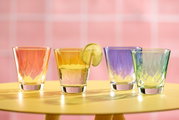 Vier bunte Trinkgläser auf einem gelben Tisch vor einem rosa Hintergrund, aus der Vorderansicht fotografiert.