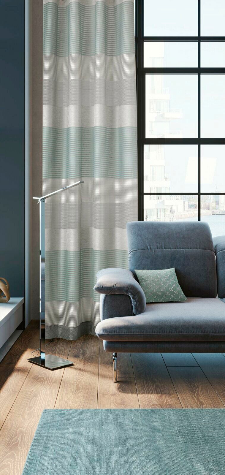 Moderner Ösenvorhang in Grau- und Grüntönen vor einem großen Fenster, neben einem grauen Sofa in einem stilvollen Wohnzimmer. Perspektive von der Seite.